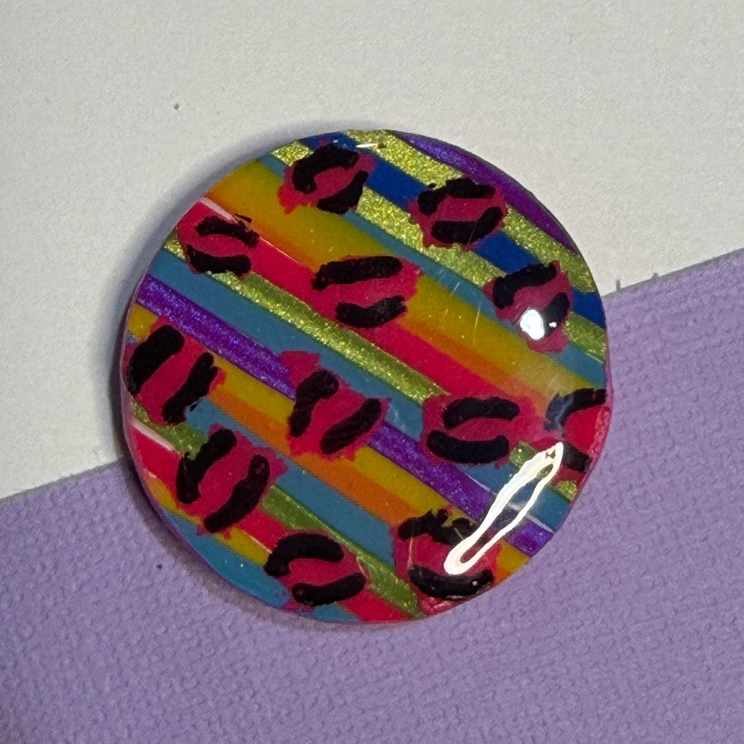 A round magnet featuring a colourful striped pattern with pink, yellow, blue, green, and purple bands overlaid with black leopard spots, resting on a split white and purple surface.