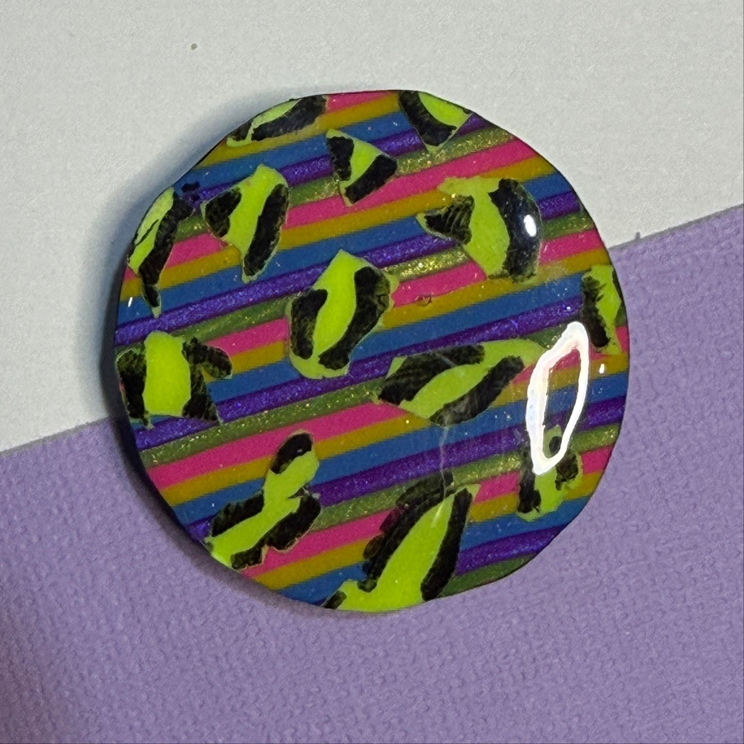 A round magnet featuring a colourful striped pattern with neon yellow-green leopard spots, resting on a split white and purple surface.