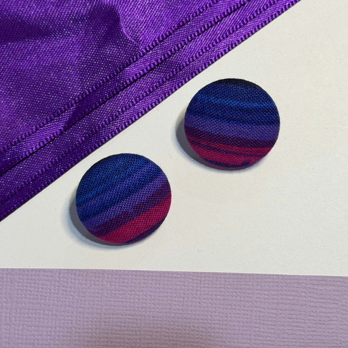 Two round fabric-covered buttons with a striped pattern of navy, purple, and magenta, displayed on a white surface next to a purple zipper.