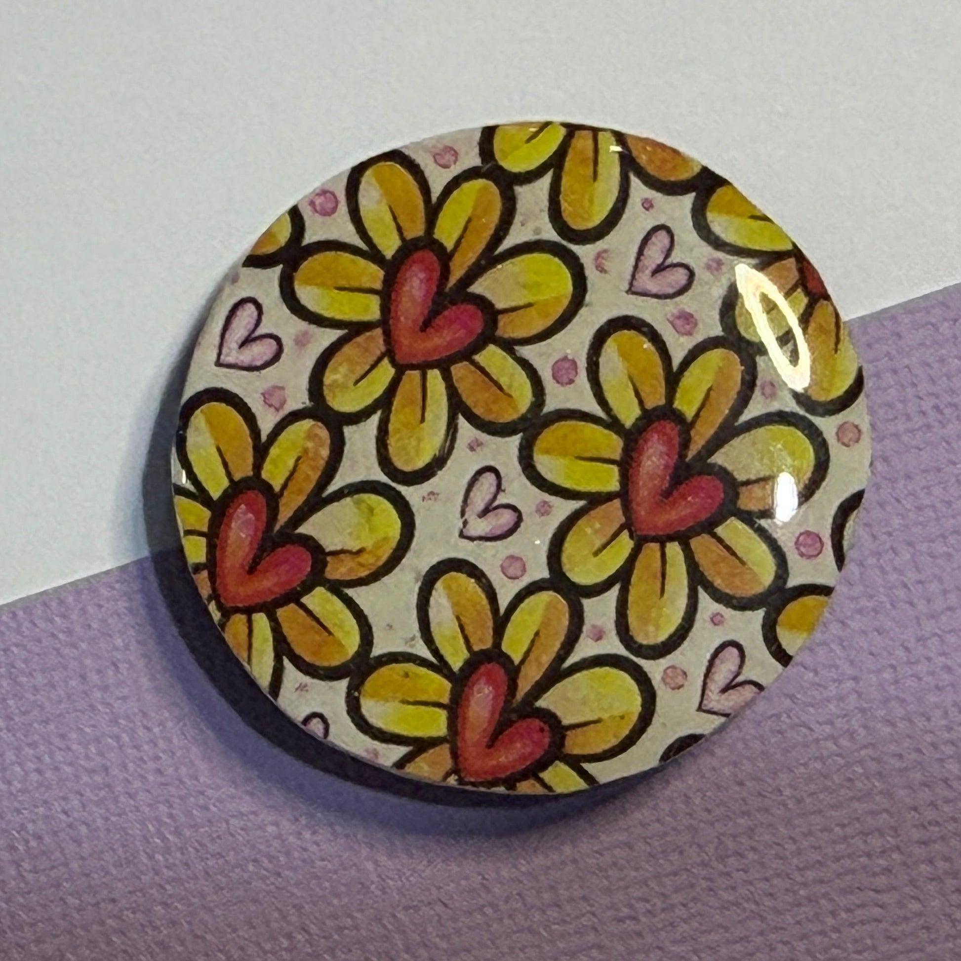 A round magnet featuring a retro-style floral pattern with yellow petals, orange-red centers, and small pink hearts on a cream background, displayed on a split purple and white surface.