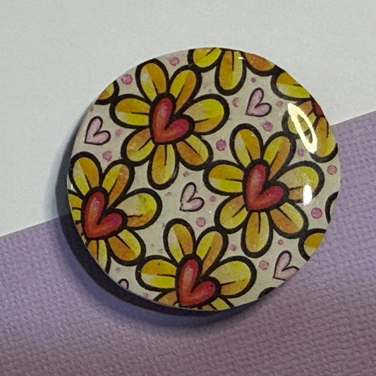 A round magnet featuring a retro-style floral pattern with yellow petals, orange-red centers, and small pink hearts on a cream background, displayed on a split purple and white surface.