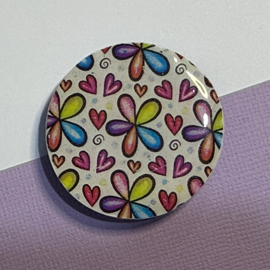 A round button badge featuring a colorful pattern of butterflies and hearts on a cream background, photographed on a split purple and white surface.