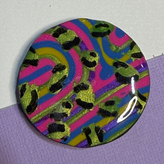 A round glossy magnet featuring a colourful abstract swirl pattern with pink, blue, yellow, and purple stripes and black leopard-like spots, photographed on a split white and lavender background.