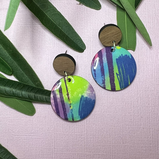 Colorful round earrings with wooden accents on a white background with green leaves.