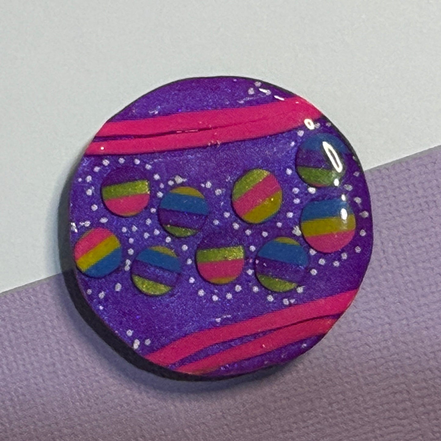 A round button or badge featuring a purple background with pink stripes, white dots, and colorful rainbow-striped circles, photographed on a split lavender and white surface.