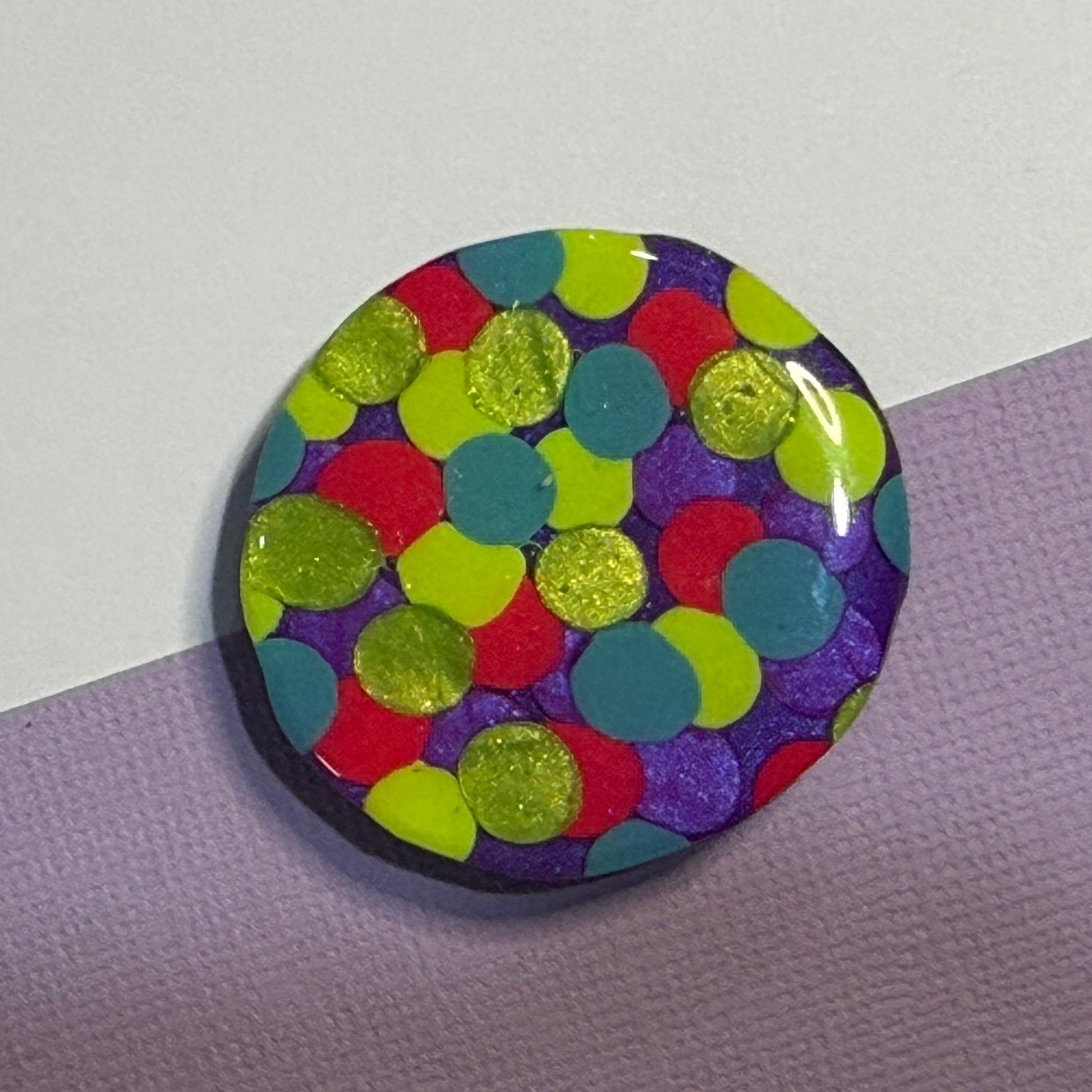 A round glossy magnet featuring a colourful confetti dot pattern with lime green, red, teal, and purple dots on a dark background, resting on a diagonally split lavender and white surface.