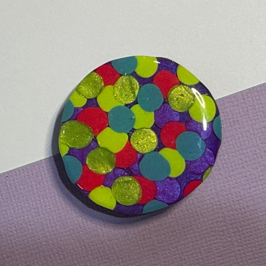 A round glossy magnet featuring a colourful confetti dot pattern with lime green, red, teal, and purple dots on a dark background, resting on a diagonally split lavender and white surface.