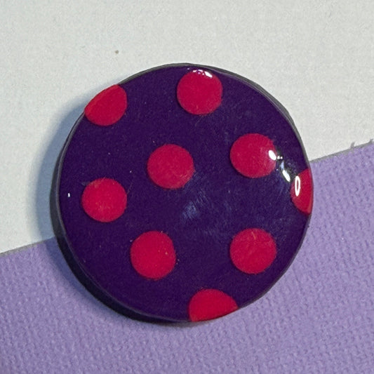 A round glossy magnet a dark purple background with pink polka dots, resting on a split white and light purple surface.