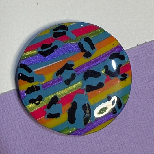 A round glossy magnet featuring a colourful striped pattern with black leopard-like spots, resting on a textured purple surface against a white background.