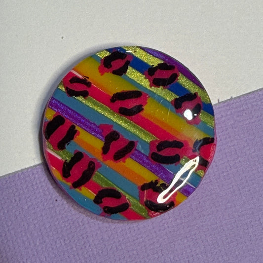 A round magnet featuring a colourful striped pattern with pink, yellow, blue, green, and purple bands overlaid with black leopard spots, resting on a split white and purple surface.