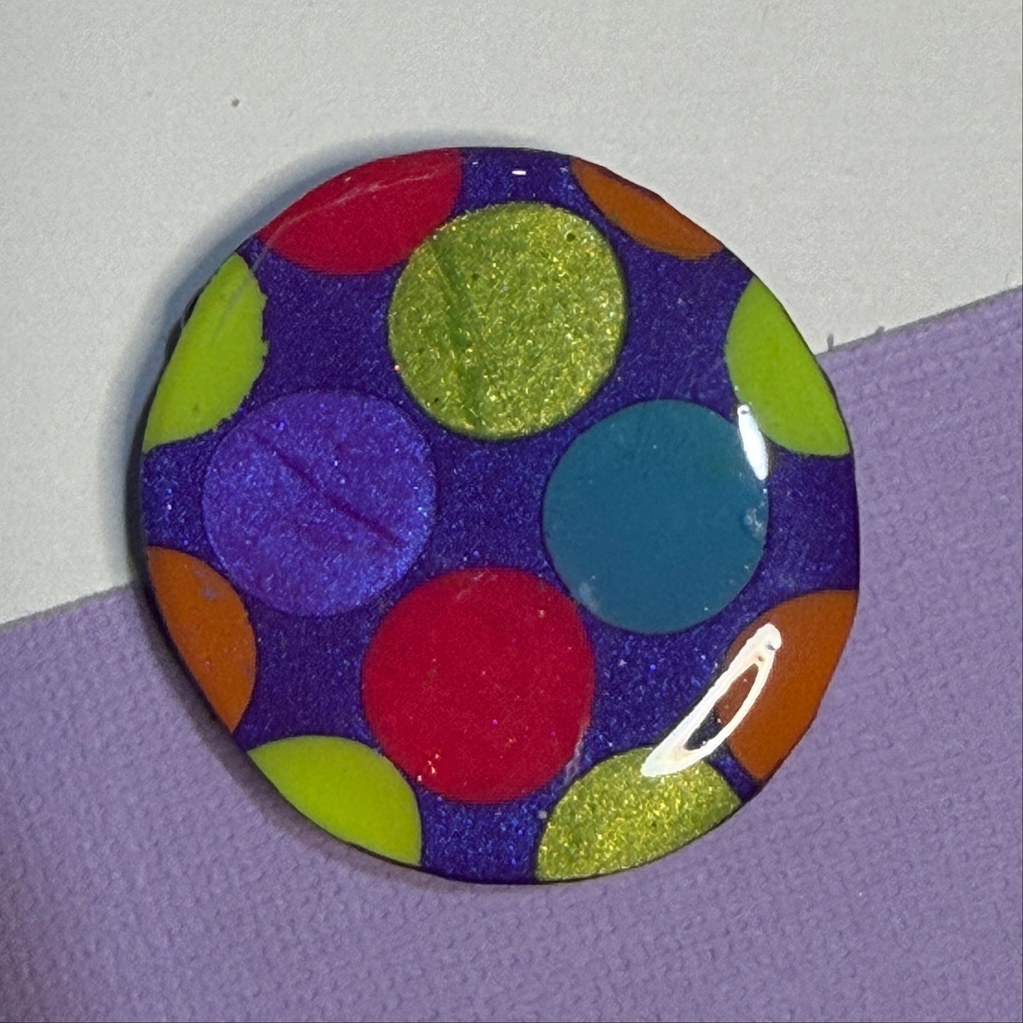 A round magnet featuring a colourful pattern of glittery and solid polka dots on a dark purple background, resting on a split white and lavender surface.