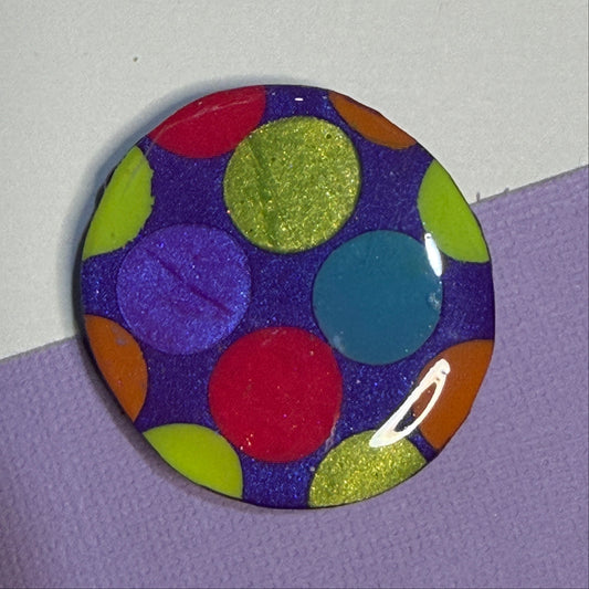 A round magnet featuring a colourful pattern of glittery and solid polka dots on a dark purple background, resting on a split white and lavender surface.