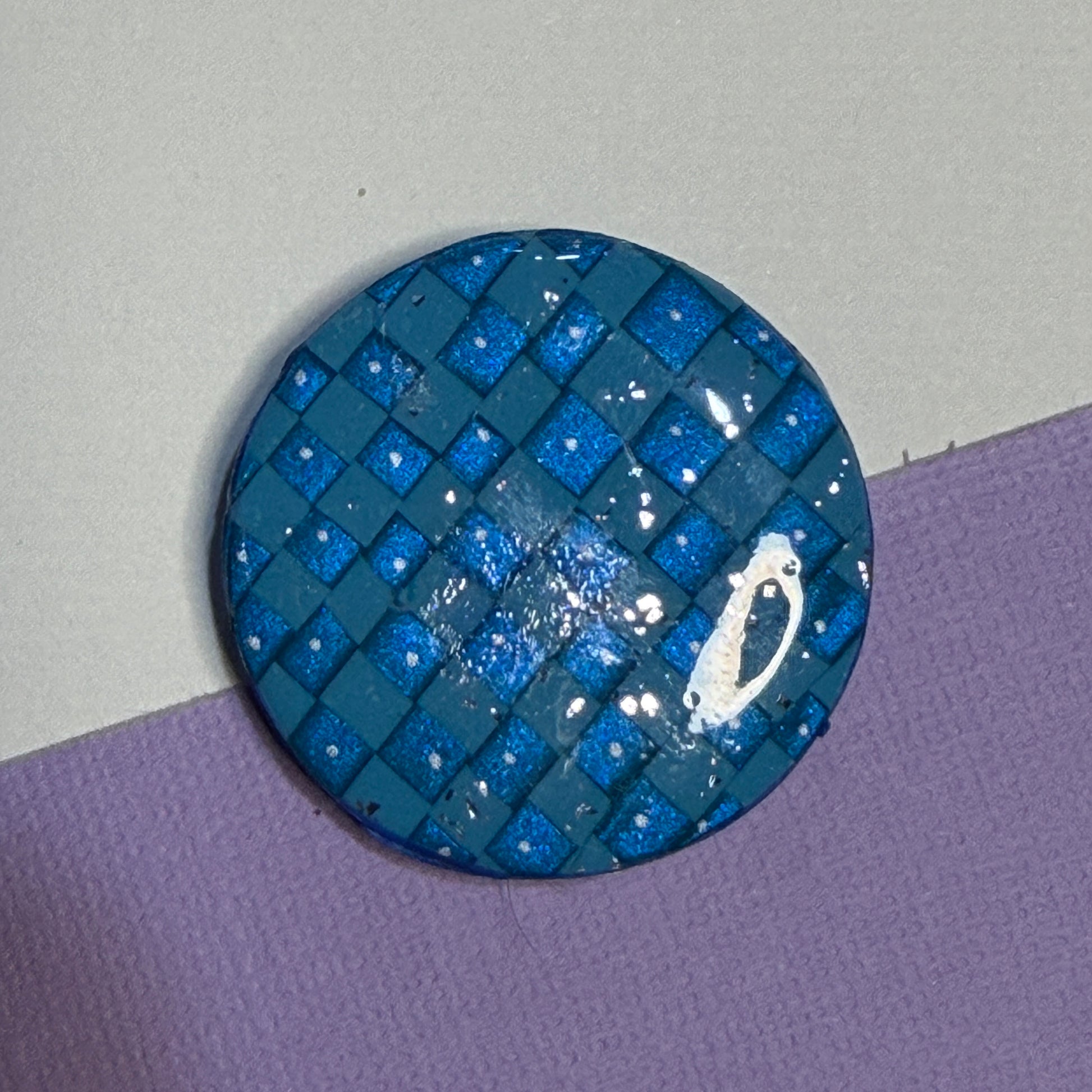 A round blue patterned magnet with a glossy finish, featuring a woven checkered design in various shades of blue with small white speckles, resting on a split white and purple background.