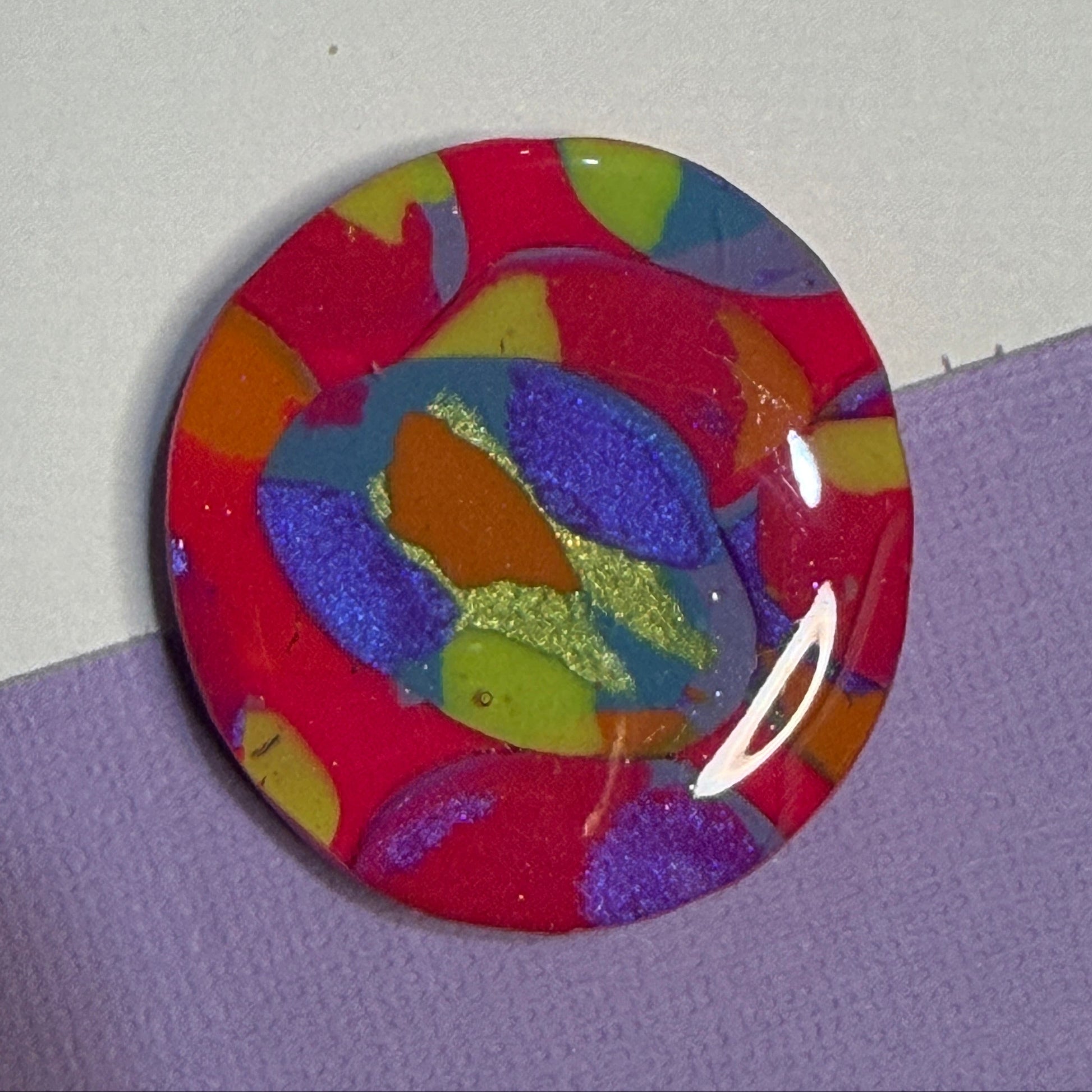 A round, glossy magnet featuring an abstract marbled pattern of red, purple, blue, yellow, and orange with glittery accents, resting on a split white and purple background.