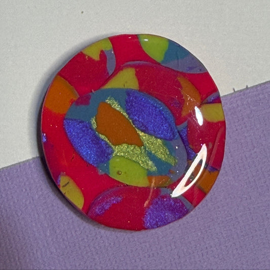 A round, glossy magnet featuring an abstract marbled pattern of red, purple, blue, yellow, and orange with glittery accents, resting on a split white and purple background.