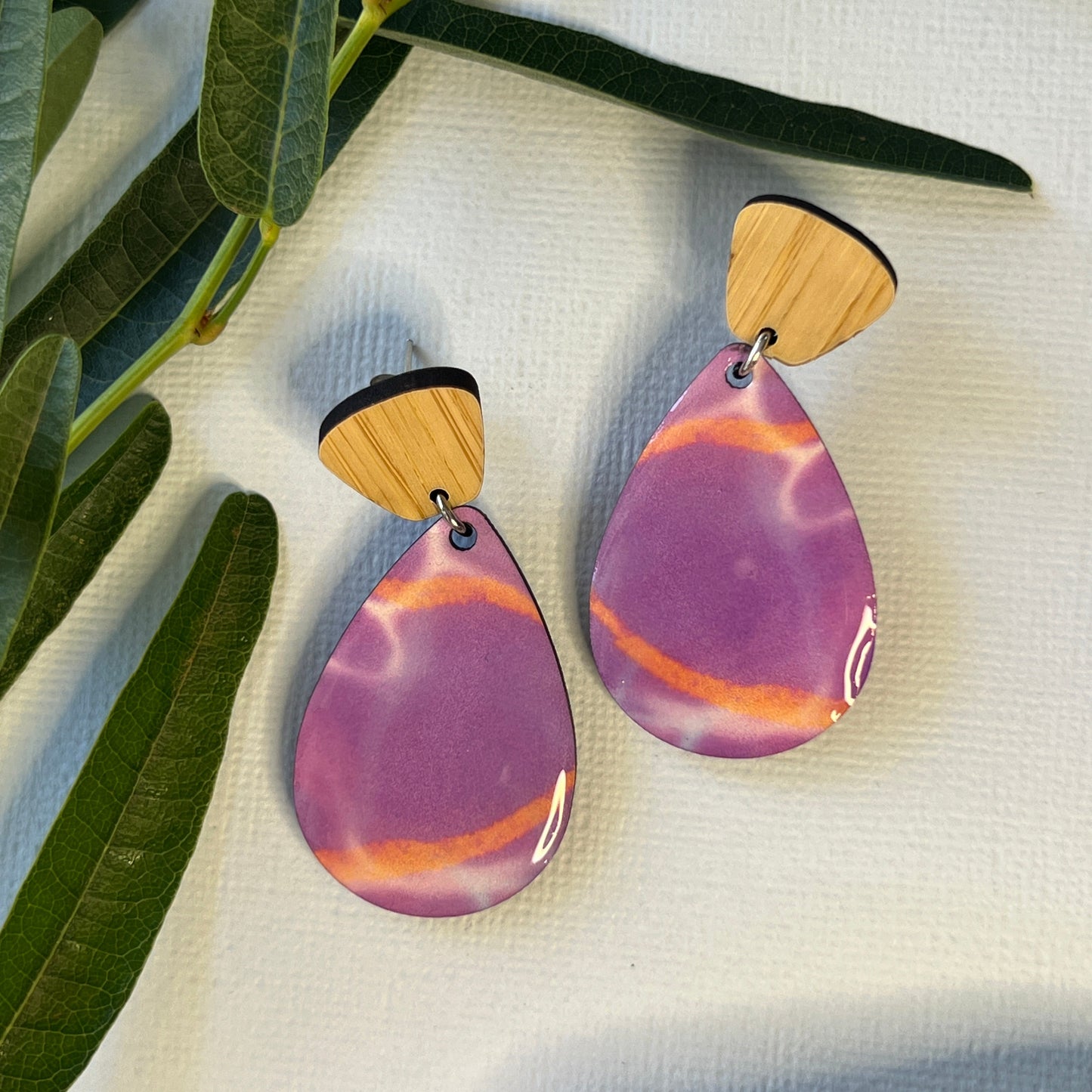 Pair of purple and gold earrings on a white fabric background with green leaves.