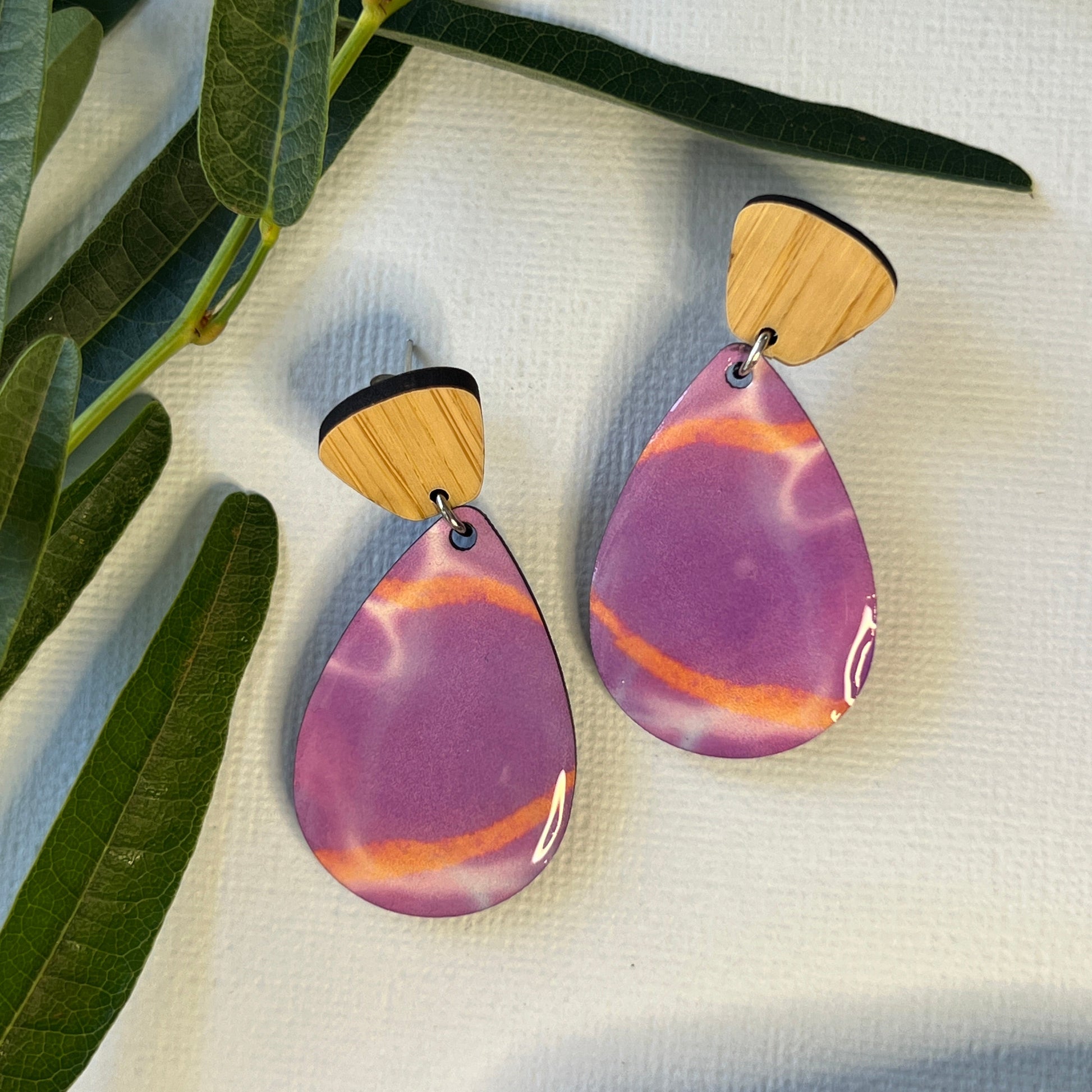 Pair of purple and gold earrings on a white fabric background with green leaves.