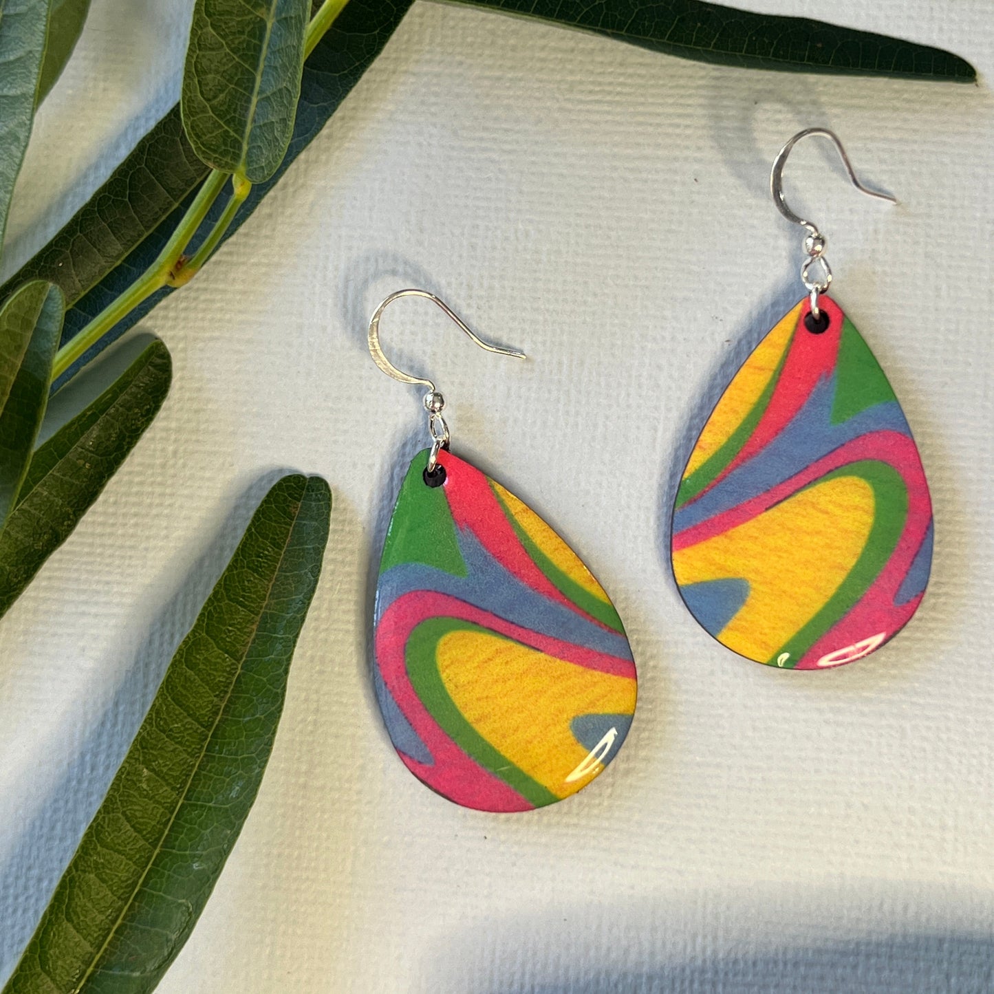 Colorful teardrop-shaped earrings on a textured surface with green leaves.